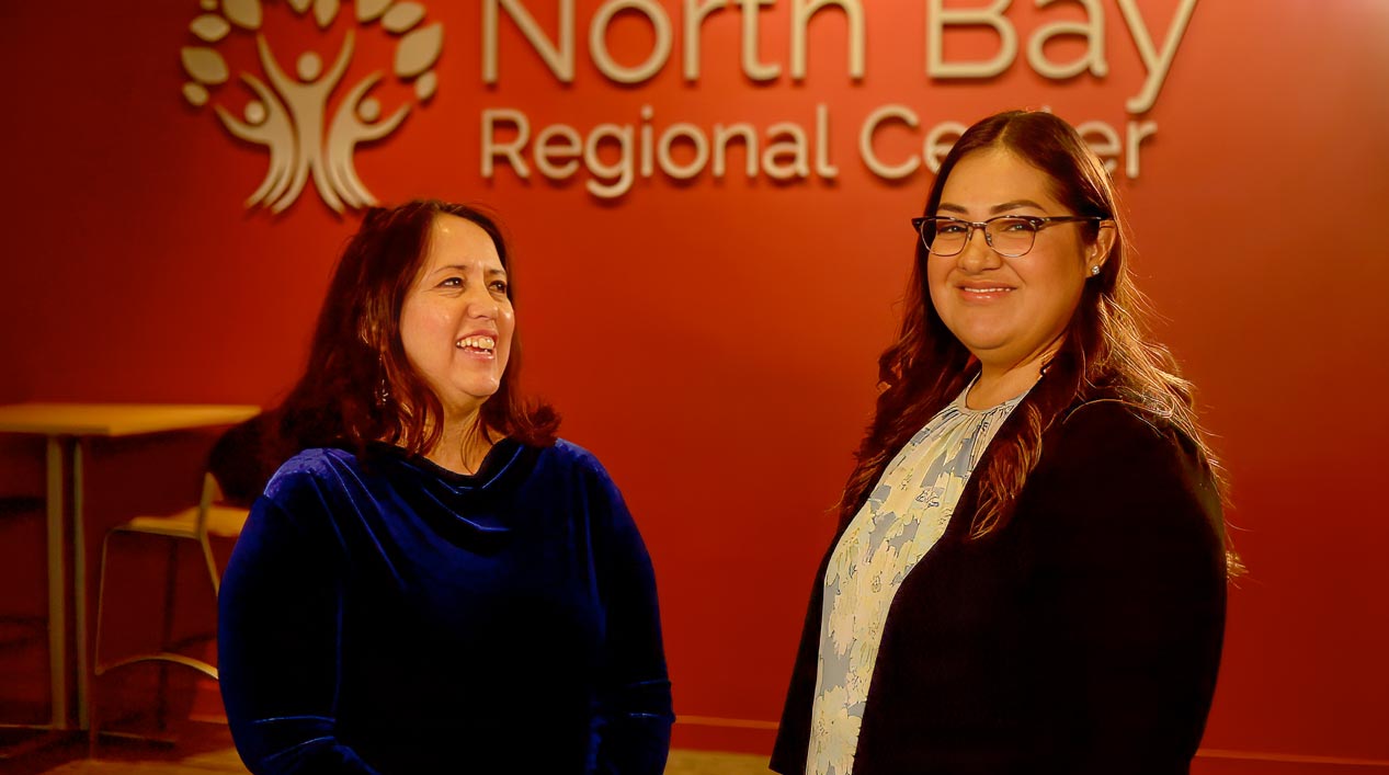 Two women smiling in conversation in front of a red wall with the text "North Bay Regional Center.