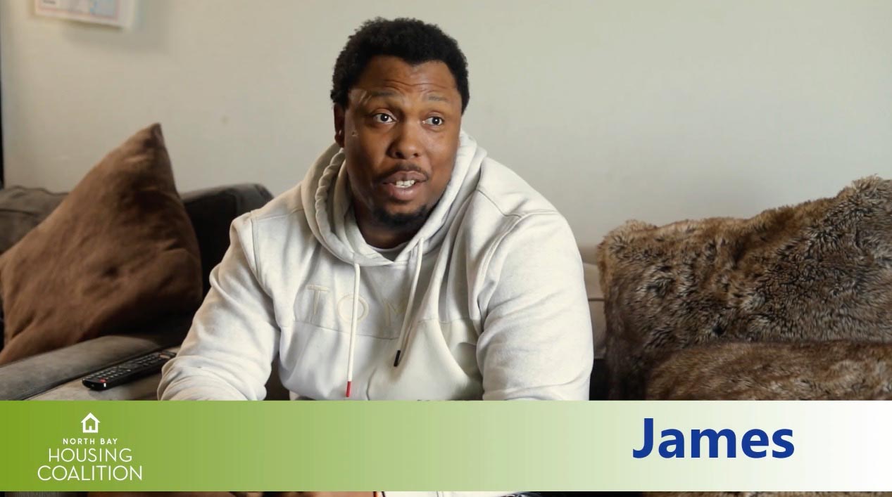 A person relaxes on a couch in a light hoodie, with the text "North Bay Housing Coalition" and "James" displayed prominently on the screen, highlighting an essential community initiative.