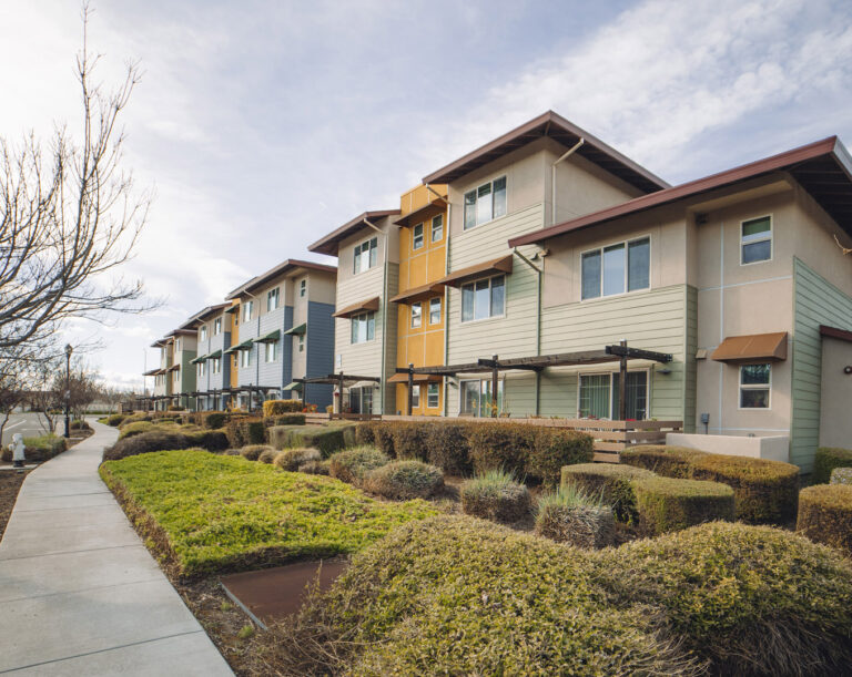 Modern three-story apartment building with beige and green siding, surrounded by neatly landscaped bushes and a sidewalk path, under a partly cloudy sky.