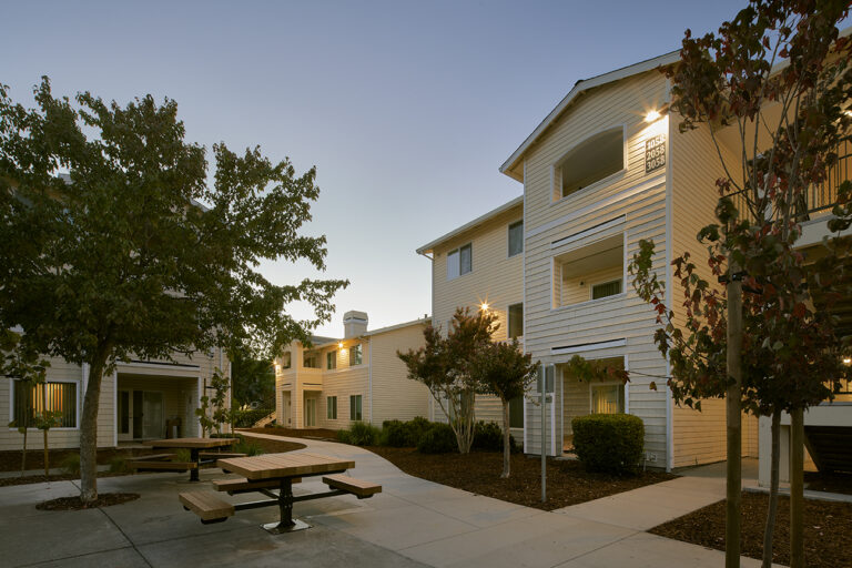 A peaceful residential complex at dusk, featuring two-story white buildings, trees, and an empty wooden picnic table in the courtyard.