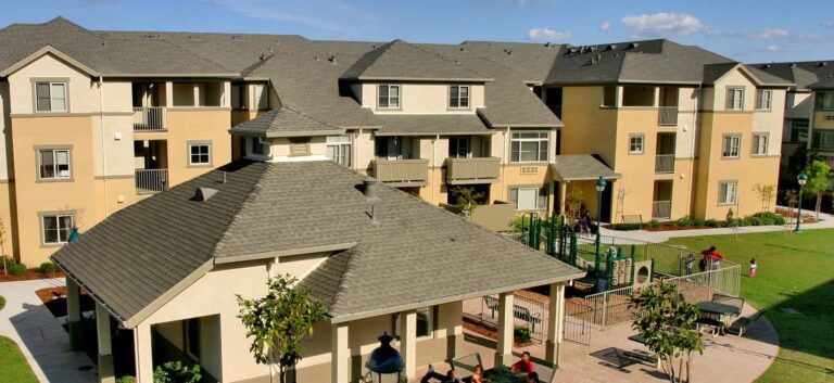 Apartment complex with three-story buildings, a playground, and a pavilion in a grassy area on a sunny day.