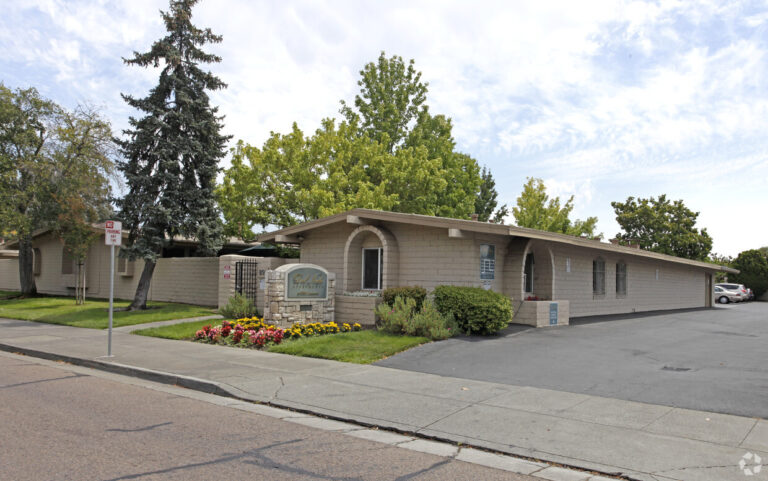 Single-story building with brick exterior and a sign for Quail Ridge Apartments, surrounded by trees and landscaping, located on a sidewalk-lined street.