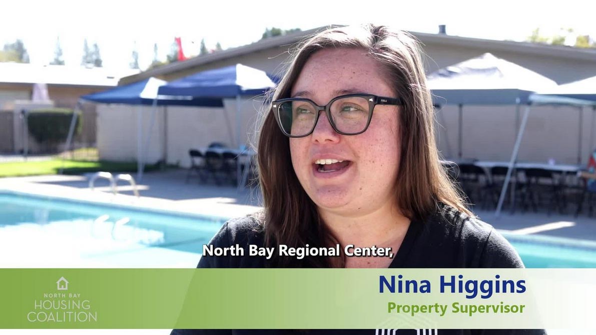 A woman wearing glasses is speaking outdoors near a swimming pool. A subtitle and a title card with her name and role are displayed.