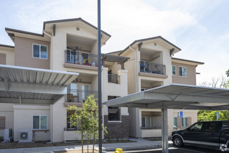 Three-story apartment building with balconies, beige siding, and covered parking in front. Small tree and vehicle visible in the foreground.